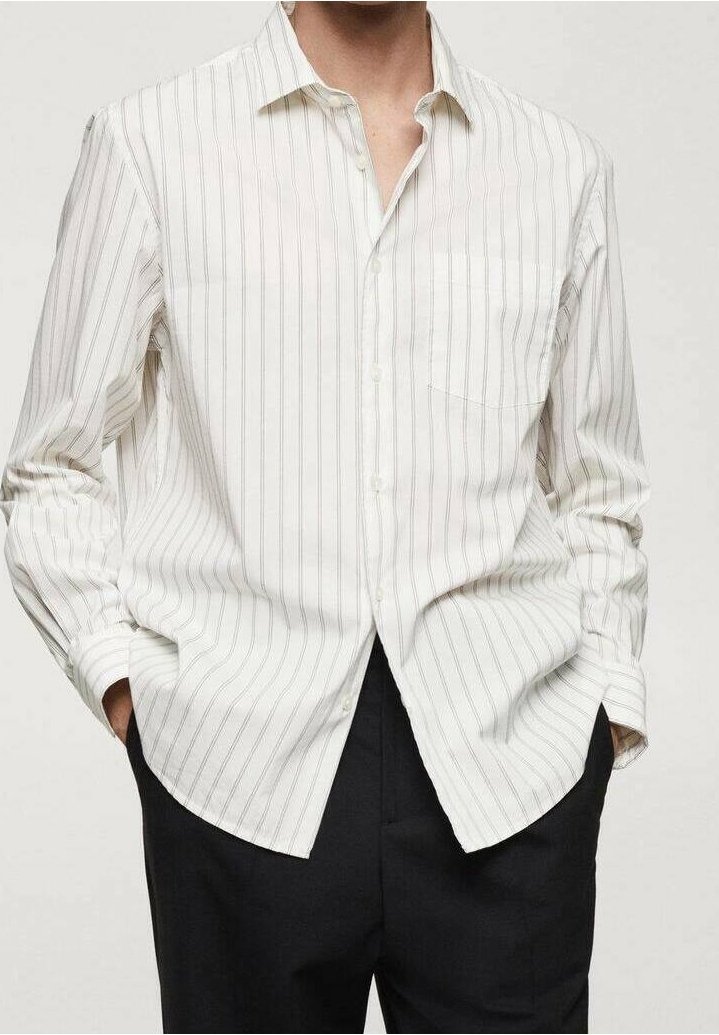 Chemise blanche à manches longues avec boutons et fines rayures verticales grises, portée non rentrée, mains dans les poches, associée à un pantalon noir.