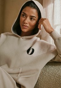 Beige hættetrøje med et stort sort broderet logo, der har snoretræk i hætten og en blød tekstur. Synlig forlommer og ribbede manchetter.