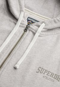 Sudadera con cremallera de color gris claro, con cordones blancos, cremallera visible y logo de "SUPERDRY ATHLETICS" impreso en el pecho.