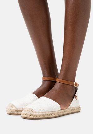 Pieds portant des espadrilles plates blanches brodées avec des lanières de cheville marron clair et des semelles en jute tressé sur un fond clair uni.