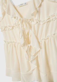 Cream-colored blouse made of lightweight fabric, featuring ruffles around the neckline and chest, with a tie closure and gathered waist.