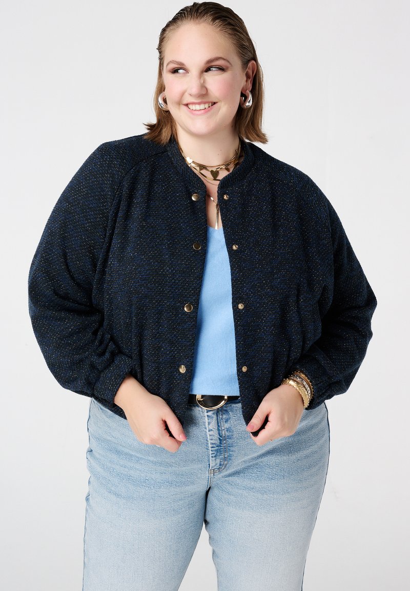 Veste bomber bleu marine texturée avec accents dorés, dotée d'un col côtelé, de boutons-pression et de poignets élastiqués, portée sur un haut bleu clair.