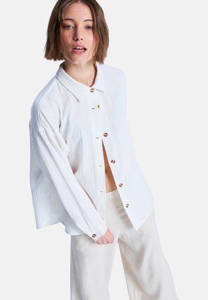 Mladá žena s krátkymi hnedom vlasmi, oblečená v nadrozmernej bielej košeli na gombíky a svetlo béžových nohaviciach, stojaca na jednoduchom pozadí.