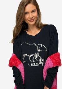Czarna koszulka z długim rękawem z graficznym szarym wzorem zwierzęcym. Modelka nosi różowy dziany szal na ramionach.