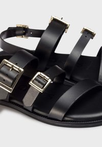 Sandalias de cuero negro con múltiples correas y hebillas en tono dorado. Textura suave y diseño de suela plana para mayor comodidad.