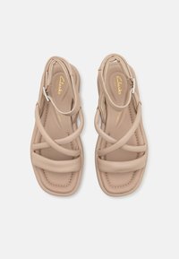 Sandali beige con cinturini, realizzati con una parte superiore sintetica liscia, cinturino alla caviglia, soletta imbottita e un design minimalista caratterizzato da linee curve e cuciture.