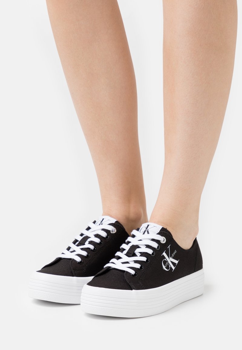 Svarta canvas sneakers med vita snören och plattformssula. Har logotyp på tungan och sidan, slät textur och rund tåkonstruktion.