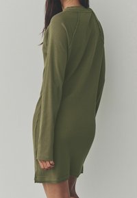 Robe longue à manches longues vert olive en tissu côtelé texturé, présentant un col rond et des accents sur les coutures latérales, se terminant à mi-cuisse.