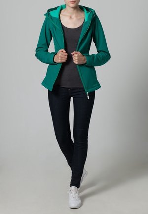 Vihreä vetoketjullinen takki, jossa on sopiva huppu, on puettu tumman paidan ja tummien farkkujen päälle. Takissa on virtaviivainen muotoilu ja sileä kankaan rakenne.