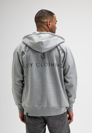 Man draagt een lichtgrijze capuchonsweater met zwarte tekst op de rug, gecombineerd met donkergrijze broek, met de rug naar de camera gekeerd.