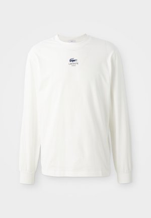 Valge pika varrukaga T-särk, mis on valmistatud puuvillast, omades ümmargust kaelust ja väikest sinist Lacoste logo rinna piirkonnas. Lihtne disain ja keha lähedane varrukate lõige.