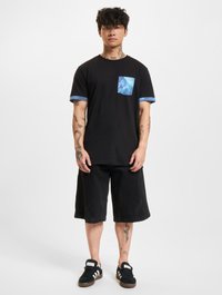 Black T-shirt with a blue patterned pocket and sleeve cuffs, paired with black shorts. Model wears black sneakers. Plain white background.