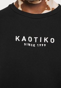 Primer plano de una persona que lleva una camiseta negra con "KAOTIKO SINCE 1999" bordado en blanco en el pecho.