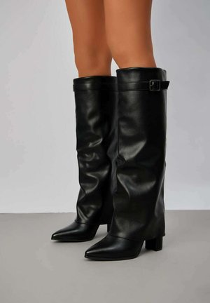 Bottes noires en cuir montantes avec un design flasque, bout pointu, talon épais et un accent de boucle sur le côté pour l'ajustement.