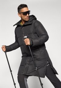 Veste rembourrée noire avec capuche, dotée d'accents zippés et de trous pour les pouces. Le modèle tient des bâtons de trekking, portant des lunettes de soleil surdimensionnées de couleur foncée.