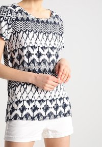 Kortärmad topp med fyrkantig halsringning, prydd med ett svart-vitt geometriskt mönster och en avslappnad passform, matchad med vita shorts.