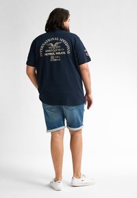 Marinblå kortärmad skjorta med gyllene text och grafikdesign på baksidan, ihopparad med ljusblå uppvikta denimshorts och vita sneakers.