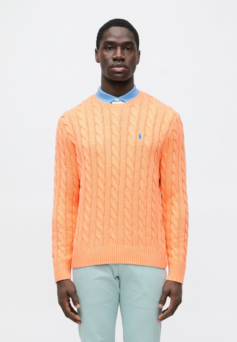 Πορτοκαλί πουλόβερ με κόμπο, με ριμπ τελείωμα, που φοράει από κάτω ένα ανοιχτό μπλε πουκάμισο με γιακά. Μικρό λογότυπο στο στήθος.