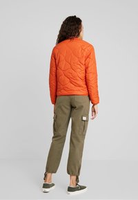 Femme de dos portant une veste matelassée orange, un pantalon cargo vert olive avec des poches, et des chaussures vertes à enfiler avec des semelles blanches.