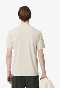 Beige kortärmad polo med krage, gjord av strukturerad tyg, sedd bakifrån. Personen bär mörkgröna byxor och håller ett ljust föremål.