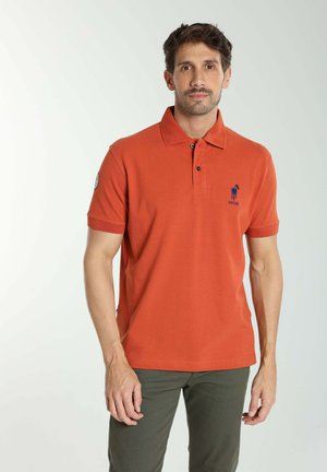 Homme portant un polo manches courtes orange avec un logo bleu brodé sur la poitrine et un pantalon vert olive, debout devant un fond uni.