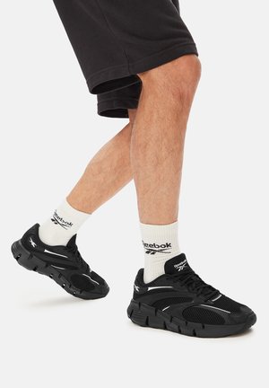 Osoba nosząca czarne sneakersy Reebok i białe skarpetki Reebok, z jedną nogą lekko uniesioną, na jednolitym jasnym tle.