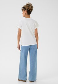 Frau mit lockigem Haar in einem Dutt, die ein schlichtes weißes T-Shirt, helle, weite Jeans und braune Schuhe trägt, steht mit dem Rücken zu einem weißen Hintergrund.