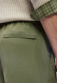 Κοντινό πλάνο ενός ατόμου που φοράει υφασμάτινο χακί παντελόνι με ετικέτα Calvin Klein και μανίκι από πλεκτό κρεμ πουλόβερ τσαλακωμένο προς τα πάνω.