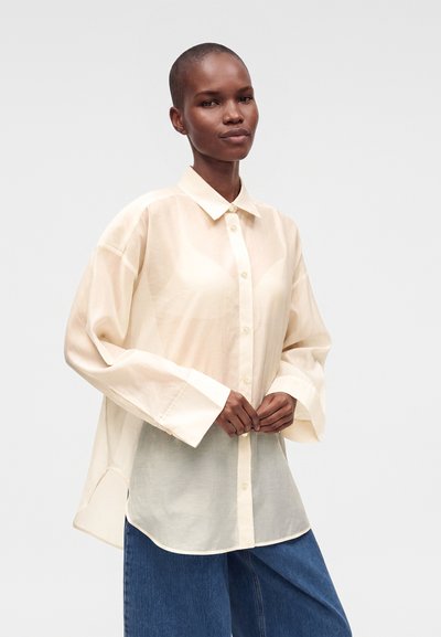 Personne aux cheveux courts portant une chemise ample et transparente couleur crème, boutonnée, et un jean bleu à jambes larges, debout les mains jointes.