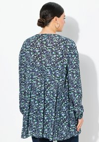 Langærmet tunika i mørkeblå stof med et blomster mønster i grøn og lyseblå. Har en plissé design bagpå og en rund halsudskæring.