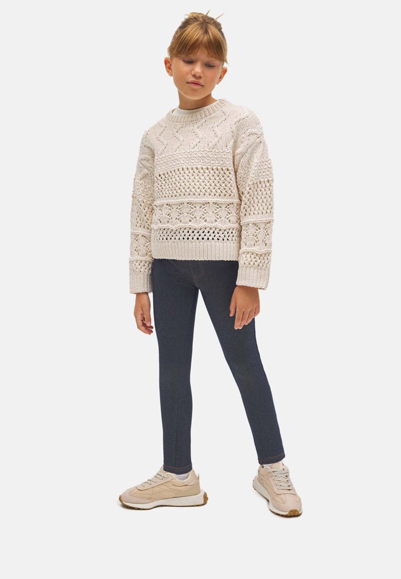 Suéter de punto en color crema con patrones texturizados, combinado con leggings oscuros y zapatillas beige. Presenta mangas largas y un escote redondo.