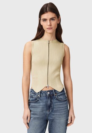 Kvinde med langt brunt hår iført ærmeløs beige ribbet top med lynlås og blå jeans med høj talje, stående foran en ensfarvet grå baggrund.