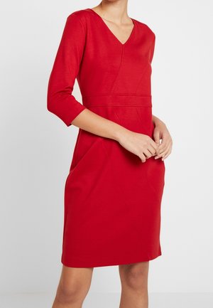 Frau trägt ein knielanges rotes Kleid mit dreiviertel Ärmeln und V-Ausschnitt, steht mit vor der Taille verschränkten Händen.