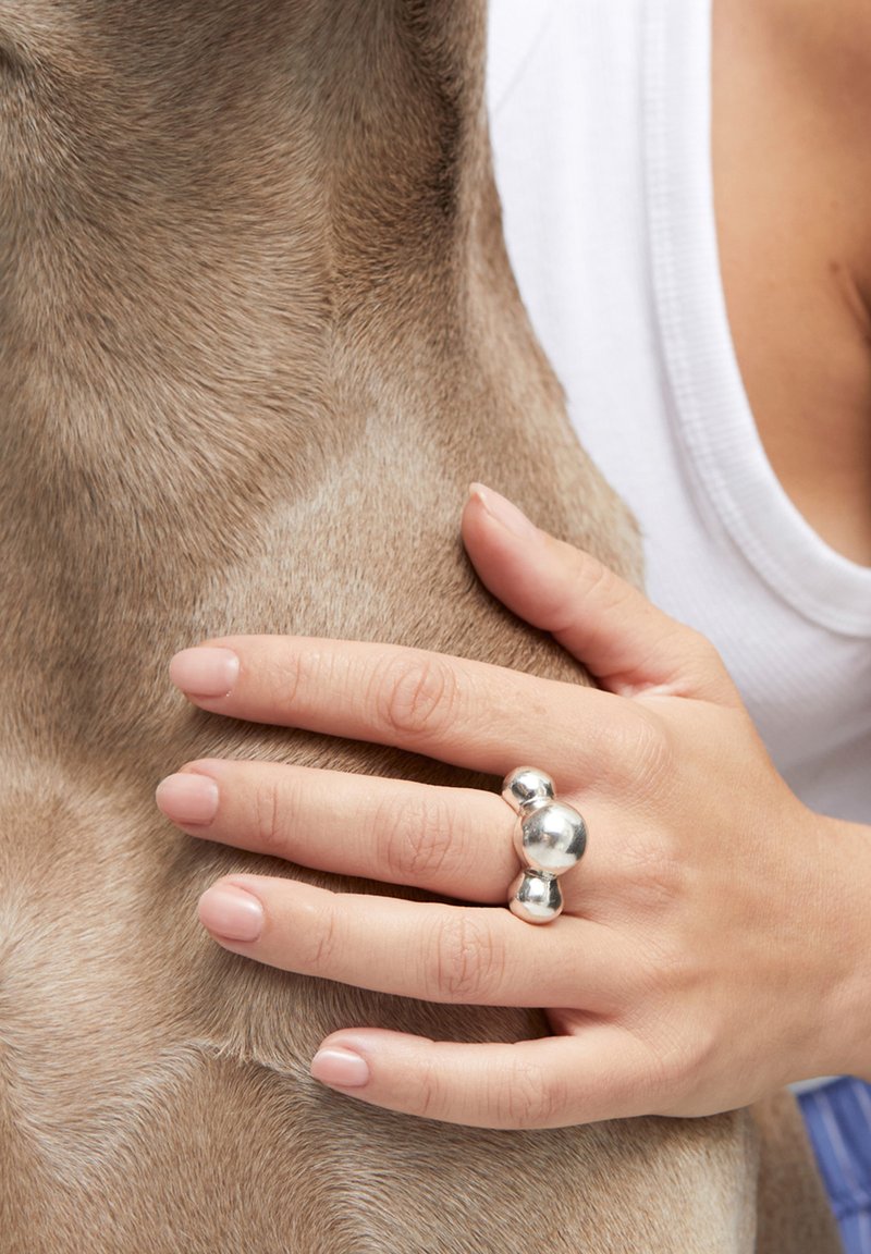 Bague en argent avec trois accents sphériques sur une main, reposant sur le pelage brun clair d'un chien. Texture lisse et design simple sont mis en avant.