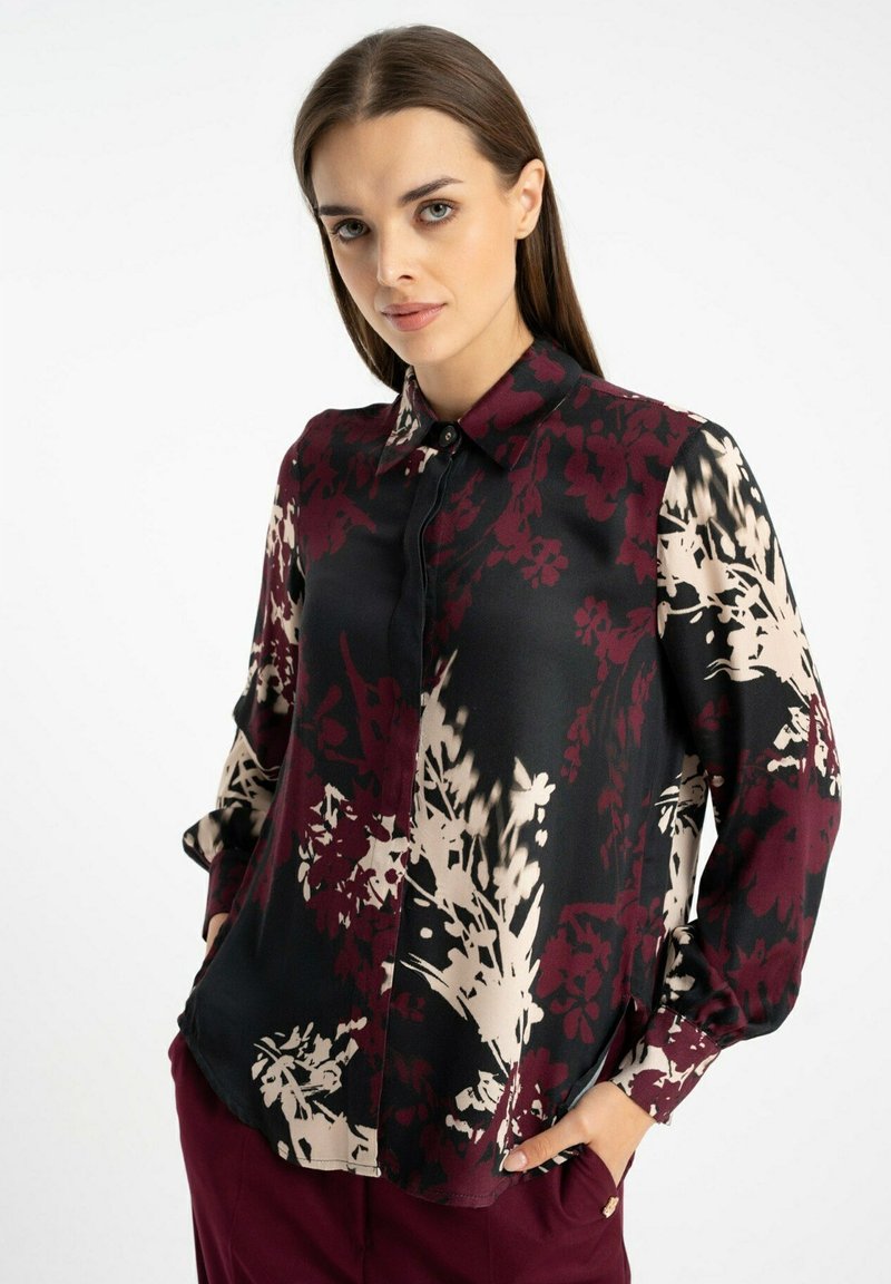 Czarna bluzka z maroonowymi i beżowymi kwiatowymi wzorami. Posiada kołnierzyk, zapięcie na guziki z przodu oraz długie rękawy. Lekko wygięty dół.