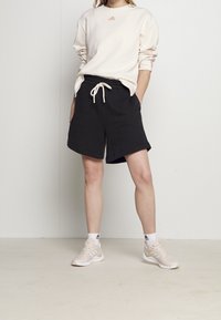 Ljusbeige sweatshirt med en liten logotyp, matchad med svarta shorts med elastisk midja och sidofickor. Ljusa skor med detaljer.