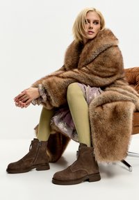 Stivali ankle in suede marrone con zip laterale, abbinati a collant a costine olive testurizzati e un cappotto marrone con bordo in pelliccia, seduti su una sedia moderna.