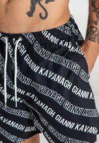 Crne kupaćice s ponavljajućim bijelim tekstom "GIANNI KAVANAGH." Elastična talija s vezicom, lagana tkanina, dizajn do koljena.
