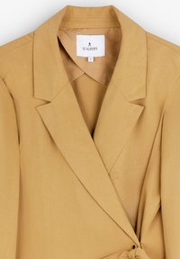 Senfgelber Blazer aus strukturiertem Stoff mit Reverskragen, Taillengürtel und einem Markenlabel innen.