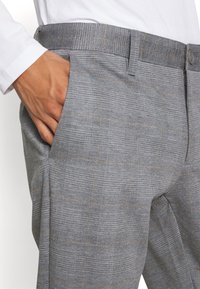 Mano in tasca di pantaloni a quadri grigi indossati con una camicia bianca a maniche lunghe.