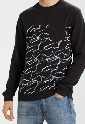 Černé tričko s dlouhým rukávem s bílým abstraktním vzorem čar, náprsní kapsa, nošeno s modrými džínami, osoba stojící s jednou rukou v kapse.