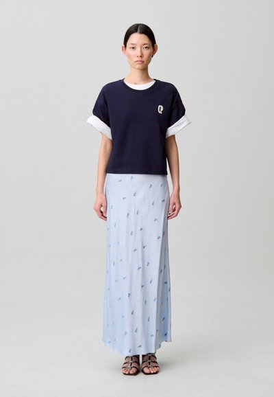 Felpa cropped blu navy con maniche arrotolate bianche, sovrapposta a una gonna lunga floreale azzurro chiaro con piccoli disegni blu. Sandali neri con cinturini.