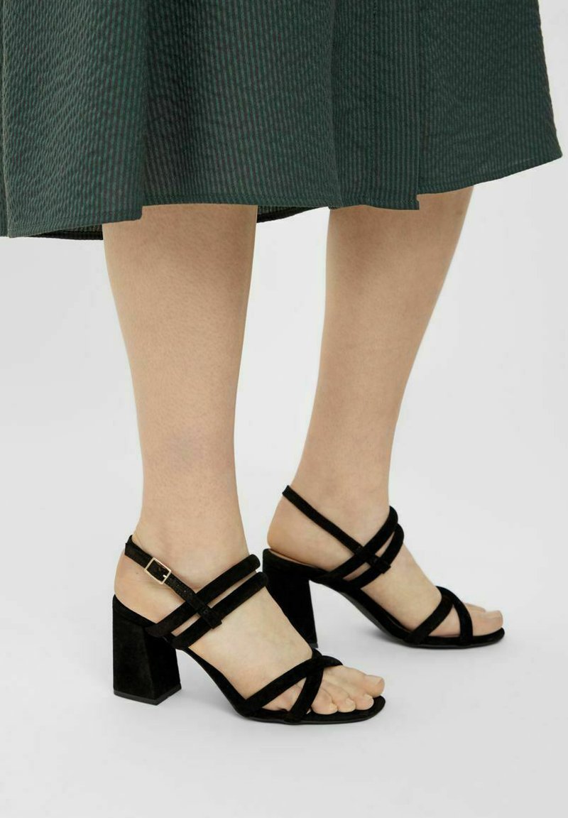 Sorte hælede sandaler med blokhæle, der har krydsede remme i blødt ruskind og en spænde lukning om anklen. Åben tå design.