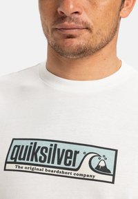 Bílý bavlněný tričko s černým a světle modrým grafickým logem s designy vlny a sopky. Kolo krk se zesíleným švem.