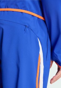 Blå idrottsbyxor med en orange accentband, vita sidostråk och en zippered ficka på vänster sida. Tillverkade av ett mjukt, elastiskt tyg.