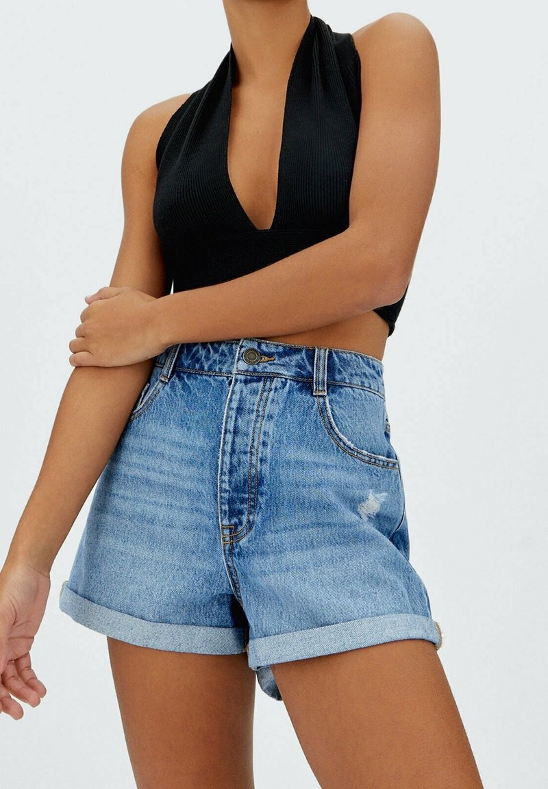 Donna con un top nero senza maniche a halter e pantaloni shorts di jeans blu a vita alta, con orlo arrotolato e un piccolo graffio.