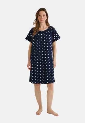 Femme debout pieds nus, portant une robe bleu marine à pois blancs, longueur genoux, à manches courtes, sur un fond uni.