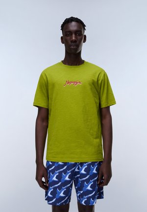 Homme portant un t-shirt Napapijri vert citron et un short bleu avec des motifs d'oiseaux blancs et bleu clair, debout devant un fond uni.
