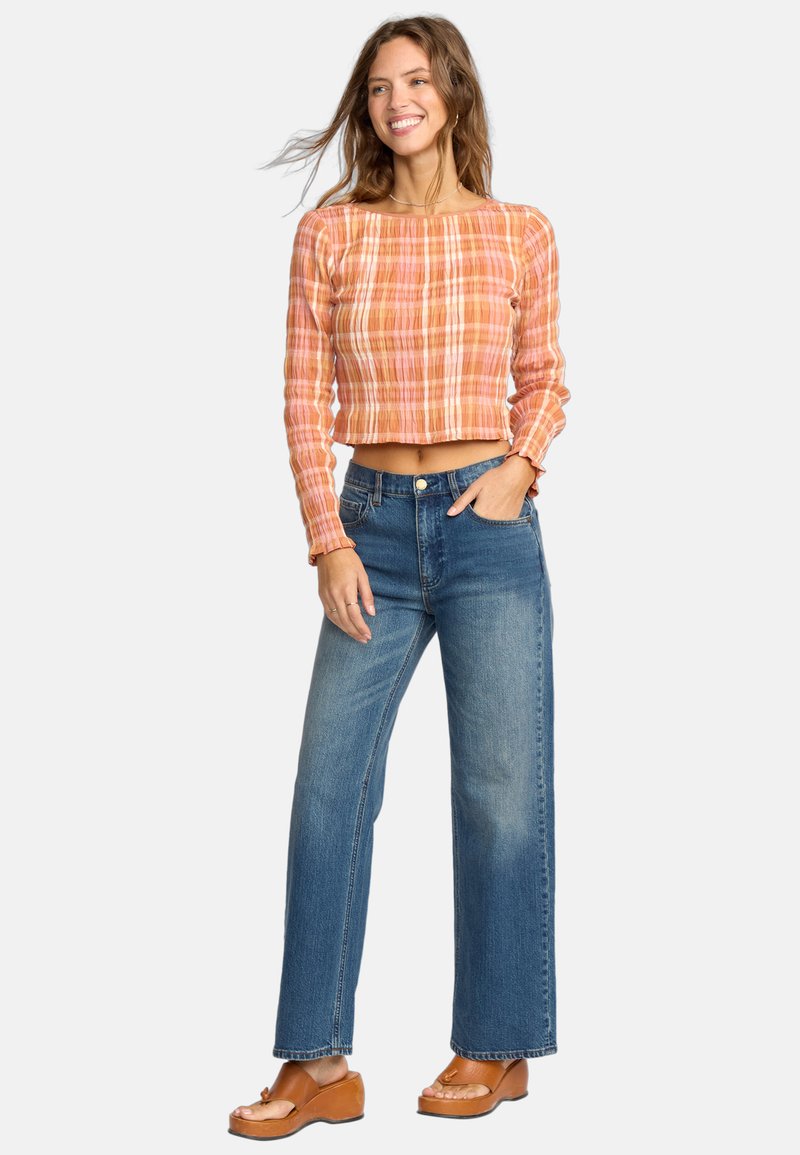 Femme souriante avec de longs cheveux portant un crop top à carreaux orange à manches longues, un jean large bleu, et des sandales à plateforme marron, se tenant avec une main dans la poche.