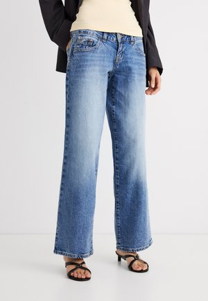 Nainen, joka pitää yhtä kättään taskussa ja pukeutuneena vaaleansinisiin leveälahkeisiin farkkuihin, mustiin korkokenkäsandaaleihin, beigen väriseen toppiin ja mustaan takkiin.